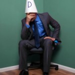 Businessman sat on a chair in the corner wearing a dunce hat