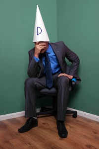 Businessman sat on a chair in the corner wearing a dunce hat